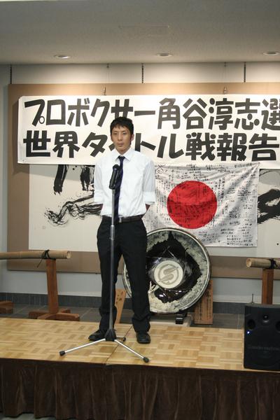 白Tシャツにネクタイを締めた角谷 淳志選手がスタンドマイクで話をしている写真